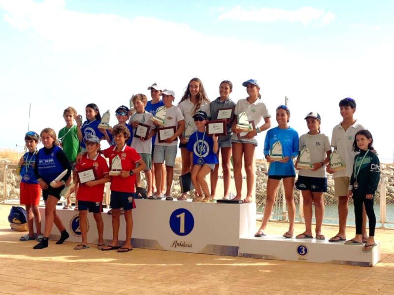 Una escena de un podio deportivo donde niños y niñas posan con sus trofeos, algunos con camisetas azules y otros con camisetas rojas. El podio tiene un número 1 en el centro.