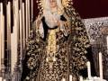 Una figura vestida en traje de gala con un manto negro y dorado, adornada con joyas y una corona. La figura está sentada en un trono de madera, rodeada por flores y velas. La escena parece ser parte de una celebración religiosa o festiva.