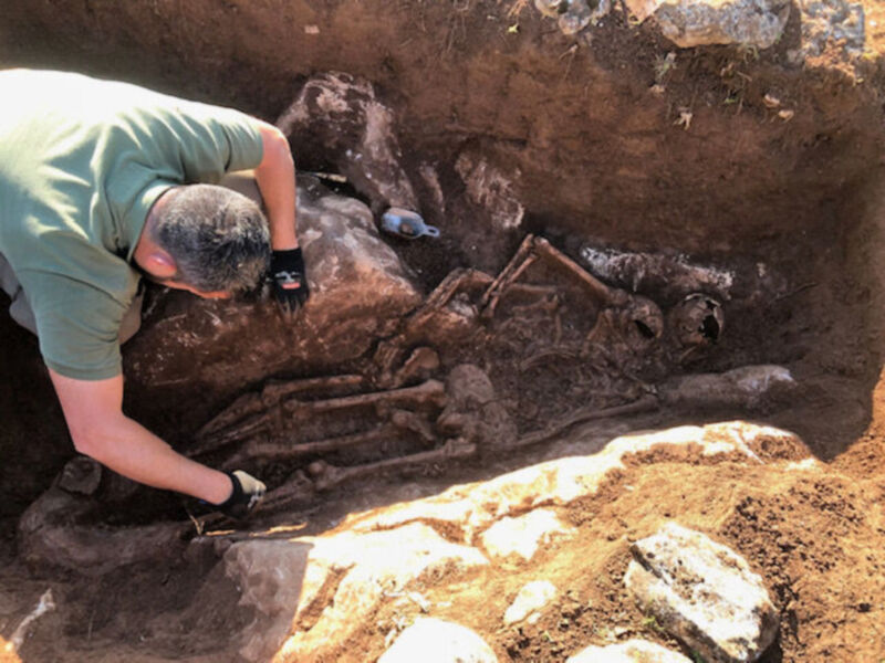 Un arqueólogo examina restos antiguos en una tumba excavada. La imagen muestra huesos y piedras, sugiriendo un entierro antiguo.