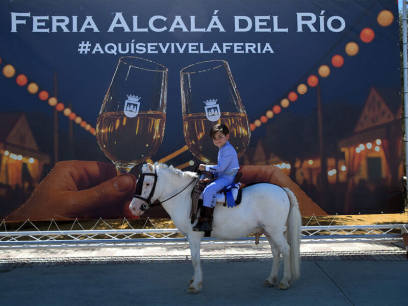 Feria Alcalá del Río, evento cultural y festivo. Imagen de un niño montando una pequeña mula frente a un cartel con el nombre de la feria y hashtags.