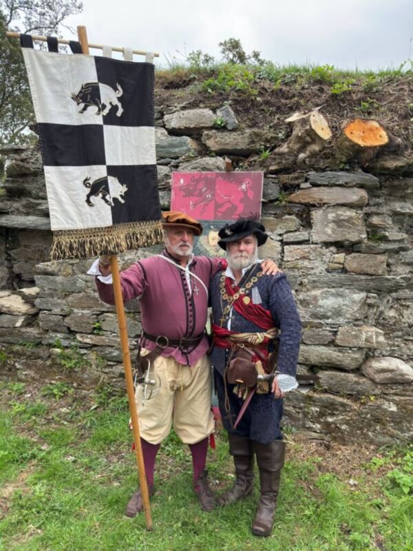 Dos hombres vestidos con trajes medievales posan junto a una bandera y un cartel en un entorno histórico.