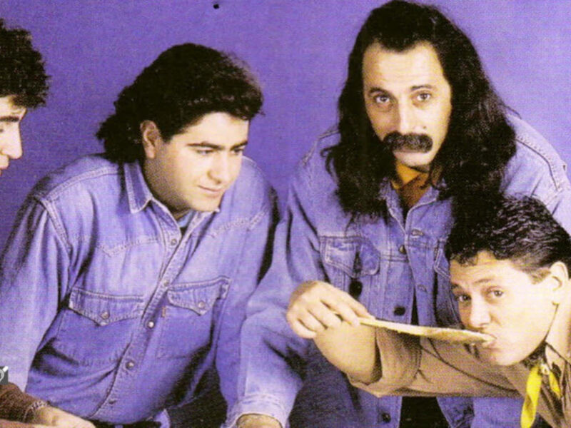 Un grupo de cuatro hombres con largas piernas, mirando hacia la izquierda. El hombre más alto tiene una barba y un bigote prominentes, mientras que los otros tres tienen cabello largo. El hombre más bajo está sosteniendo algo en sus manos. Todos están vestidos con camisas azules y pantalones oscuros. El fondo es un color azul claro.