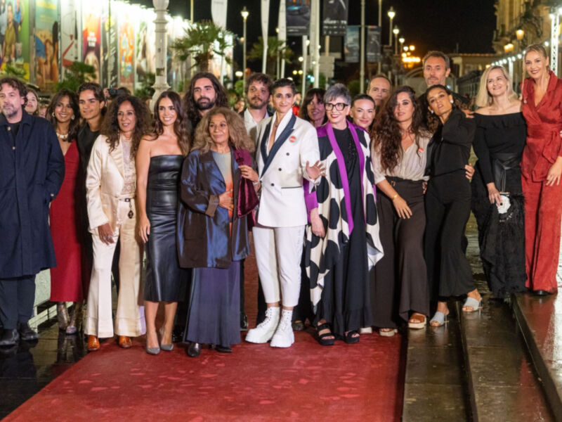 Una multitud de personas posan en un escenario nocturno, vestidas con elegantes trajes y vestidos de colores vibrantes. La escena está iluminada por faroles y carteles en el fondo, creando un ambiente festivo.