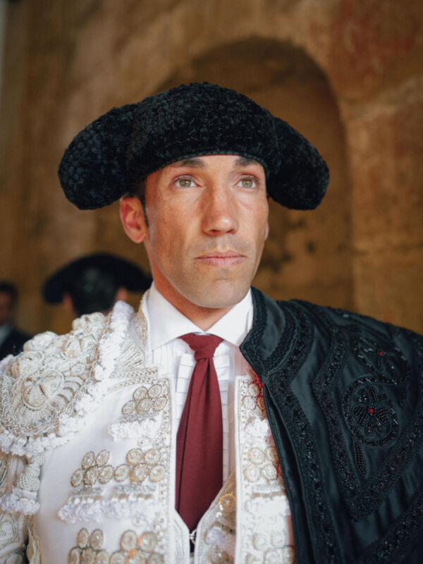 Un hombre vestido con un traje tradicional español, incluyendo una chaqueta de terciopelo blanco con detalles bordados y un sombrero negro. La escena se desarrolla en una arquitectura de estilo colonial con un fondo de paredes de ladrillo.