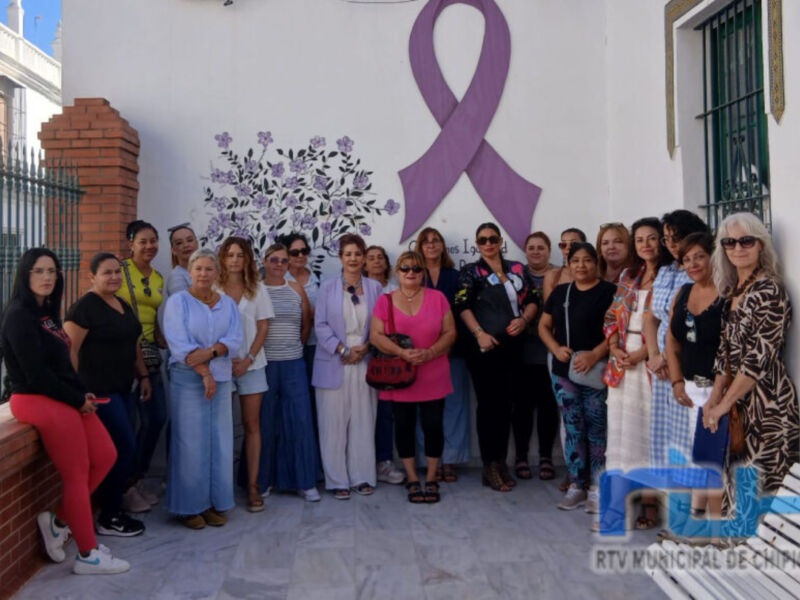 Una imagen de un grupo de personas posando frente a una pared con un lazo morado y flores, probablemente en apoyo o concienciación sobre una causa específica. La imagen está acompañada por el logo de RTV Municipal de Chiclona en la parte inferior derecha.