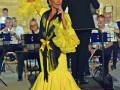 Una mujer en un traje de flamenca amarillo y negro, con un velo azul en la cabeza, canta frente a una orquesta. El escenario es al aire libre con un muro de piedra en el fondo y una placa roja que dice "Municipal de Sevilla".