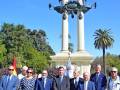 Un grupo de personas en trajes formales se encuentra frente a una estructura monumental con columnas y un escudo en el cielo azul.
