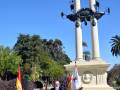 Un hombre habla en un micrófono frente a una estatua de dos leones sobre columnas, con banderas españolas y otras en el fondo. El cielo es azul claro y hay árboles verdes alrededor.