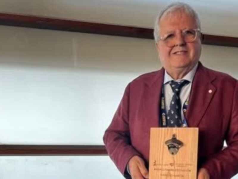 Un hombre en traje rojo sostiene un trofeo con una figura de una botella. La imagen parece ser de un evento o ceremonia formal, posiblemente un premio en un congreso o conferencia.