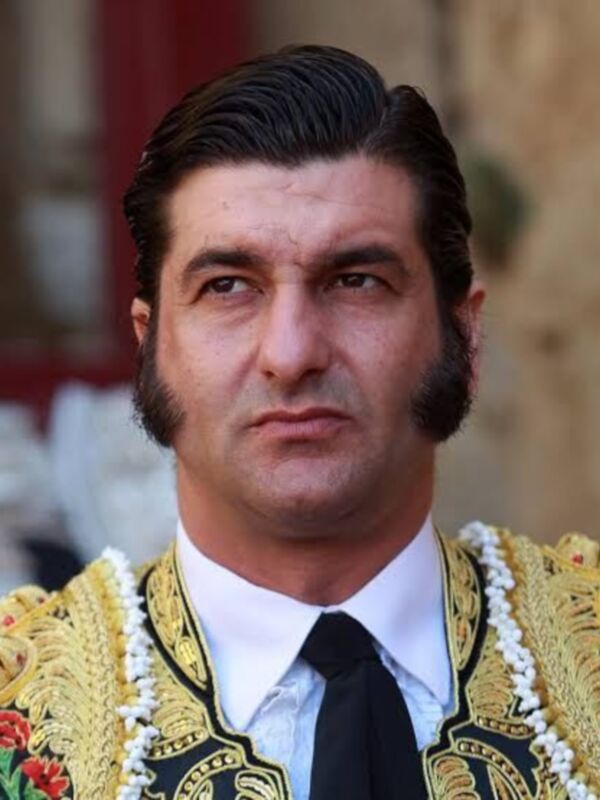 Retrato de un torero con traje tradicional, incluyendo corbata y chaqueta bordada. El fondo muestra un ambiente histórico o ceremonial, posiblemente dentro de una plaza taurina.