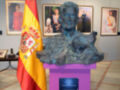 Busto de un político en una estatua, con una bandera española y varias fotos de personas en el fondo.
