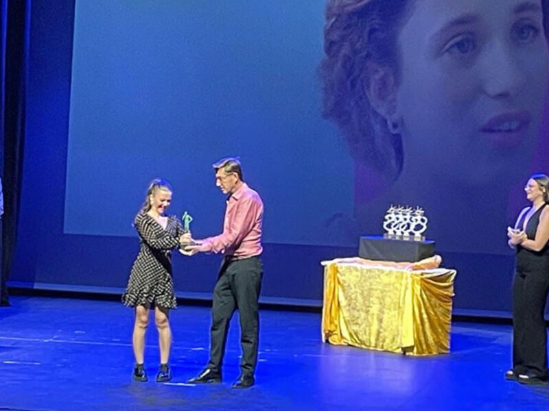 Una mujer recibe un premio de manos de un hombre en una ceremonia de reconocimiento. La escena se desarrolla sobre una gran pantalla azul con un rostro en la parte superior. La mujer sostiene el trofeo mientras sonríe, y el hombre a su lado parece emocionado. La iluminación es brillante, enfocándose en los dos protagonistas de la escena.