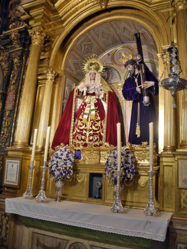 Altar de una iglesia con una imagen de la Virgen María vestida en rojo y dorado, rodeada por velas y flores. Arriba hay un arco dorado con detalles geométricos. A la derecha, una figura vestida en azul con un sombrero y un bastón. El entorno es dorado y ornamental, con detalles de madera y cristal.