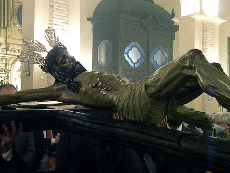 Una imagen de una estatua de Cristo crucificado en una iglesia, rodeada por personas que la sostienen. La estatua muestra el rostro de Cristo con una corona de espinas y un manto verde. La iglesia tiene vitrales azules con formas geométricas y una torre en el fondo.