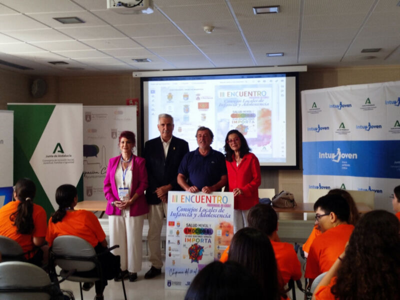 Presentación en un evento empresarial, con personas de diferentes edades y roles. En el centro hay una pantalla proyectando un slide titulado "REENCUENTRO". A la izquierda, hay una persona con una chaqueta rosa y a la derecha, una persona con una camiseta roja. En el fondo hay varios banners de empresas y organizaciones, incluyendo "IntuJoven" y "A". La sala tiene iluminación de techo y asientos para el público.