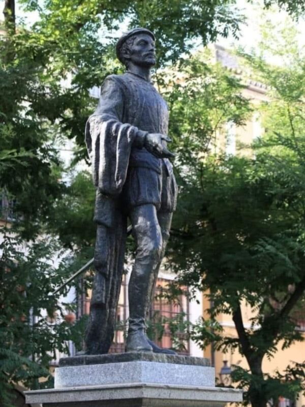 Una estatua de un hombre vestido con traje medieval, posiblemente un personaje histórico o literario. Se encuentra en un entorno urbano con árboles y edificios al fondo, lo que sugiere un parque o plaza. La estatua es de piedra y presenta detalles finos en la ropa y el sombrero.