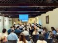 Una sala de conferencias con público sentado en sillas. La pantalla grande muestra información, y hay una persona al frente hablando. El techo tiene vigas de madera y la pared está decorada con cuadros.