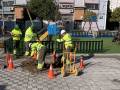 Trabajadores en uniforme amarillo realizando mantenimiento en una calle, con cascos y herramientas. Conos de seguridad alrededor del agujero excavado en el asfalto, cerca de una zona infantil y edificios.